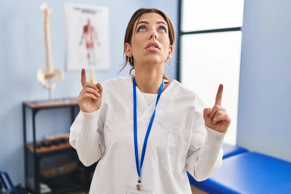 Молодая Брюнетка Работающая Клинике Поражена Удивлена Глядя Вверх Показывая Пальцами — стоковое фото
