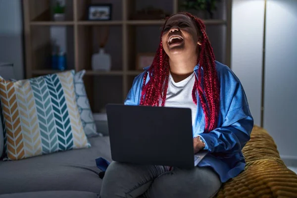 African american woman with braided hair using computer laptop at night angry and mad screaming frustrated and furious, shouting with anger. rage and aggressive concept.
