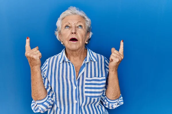 一位白发苍苍的老妇人站在蓝色的背景上 惊讶地抬起头 用手指和胳膊指指着 — 图库照片