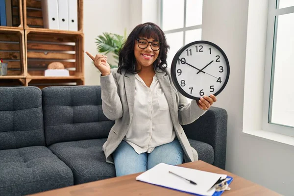 Hispanic woman working at therapy office holding clock smiling happy pointing with hand and finger to the side