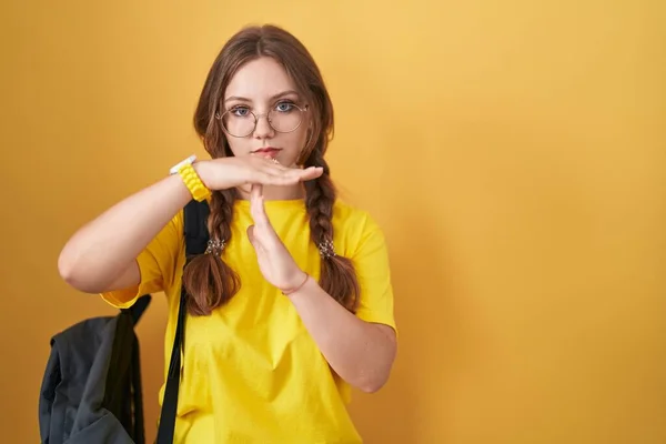 Young caucasian woman wearing student backpack over yellow background doing time out gesture with hands, frustrated and serious face