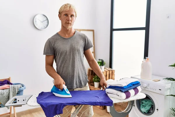 Young Blond Man Ironing Clothes Laundry Room Thinking Attitude Sober — Stock Photo, Image