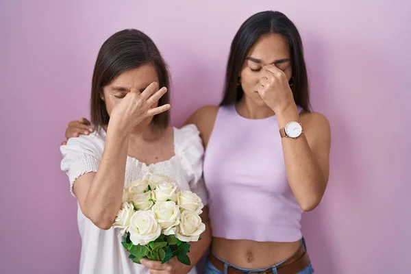 Hispanic mother and daughter holding bouquet of white flowers tired rubbing nose and eyes feeling fatigue and headache. stress and frustration concept.