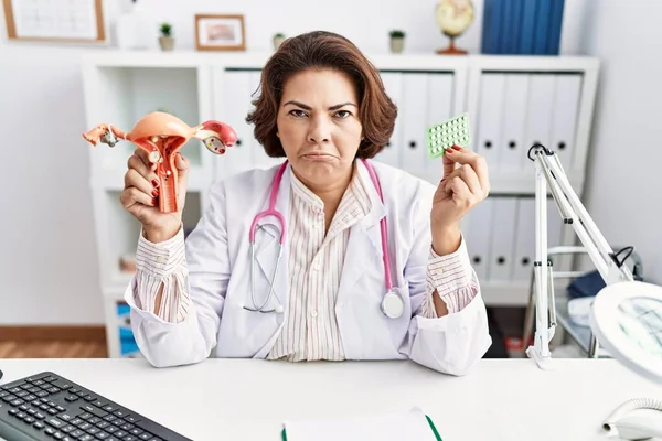 Middle age hispanic doctor woman holding anatomical female genital organ and birth control pills skeptic and nervous, frowning upset because of problem. negative person.