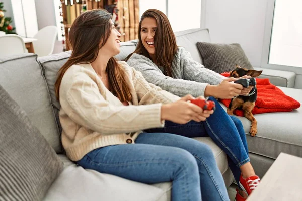 Two women playing video game sitting with dog by christmas decor at home