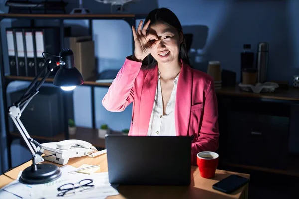 Chinese young woman working at the office at night doing ok gesture with hand smiling, eye looking through fingers with happy face.