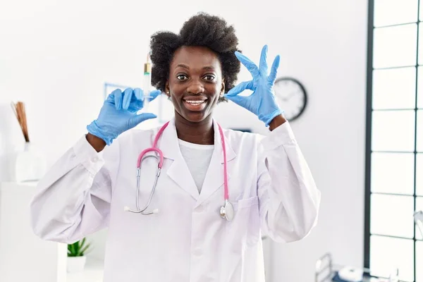 African doctor woman holding syringe at medical clinic doing ok sign with fingers, smiling friendly gesturing excellent symbol