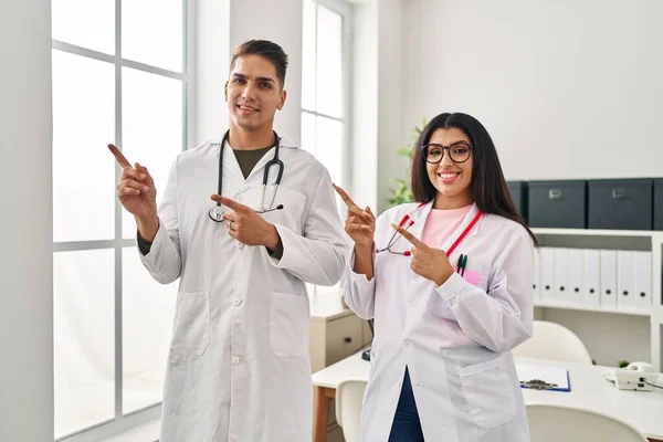 Young doctors wearing uniform and stethoscope at the clinic smiling and looking at the camera pointing with two hands and fingers to the side.