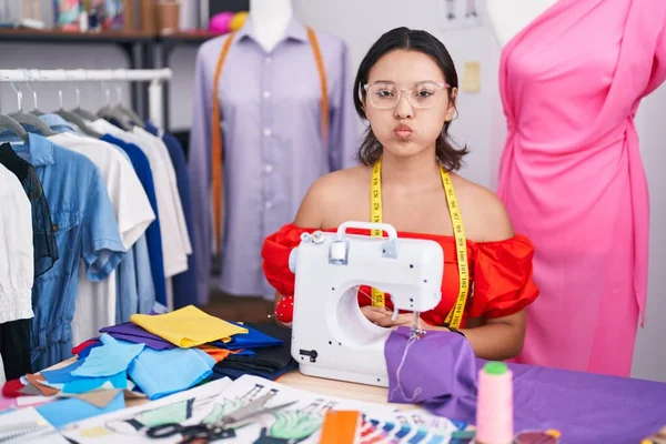 Hispanic young woman dressmaker designer using sewing machine puffing cheeks with funny face. mouth inflated with air, crazy expression.