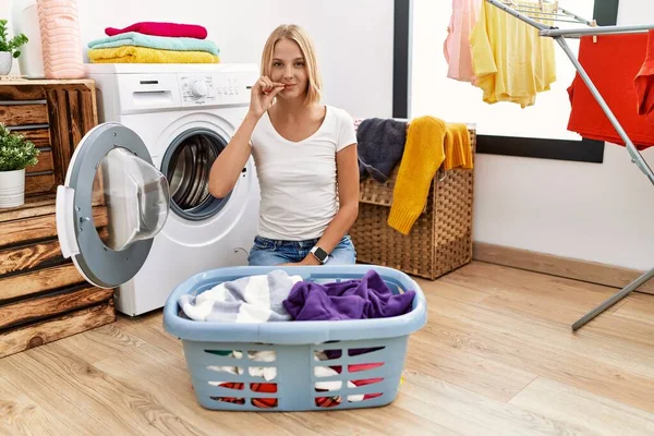 Young caucasian woman doing laundry with clothes in the basket mouth and lips shut as zip with fingers. secret and silent, taboo talking