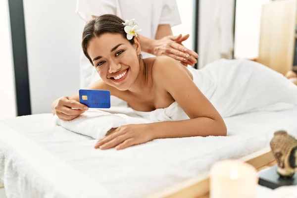 Young latin woman relaxed having back massage holding credit card at beauty center