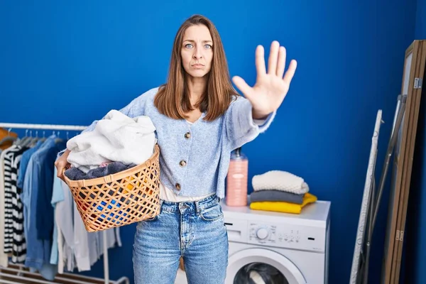 Young woman holding laundry basket with open hand doing stop sign with serious and confident expression, defense gesture