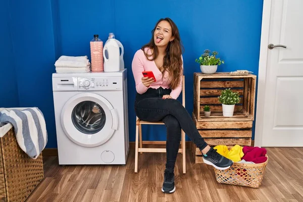 Young Hispanic Woman Sitting Waiting Laundry Using Smartphone Sticking Tongue — Stockfoto