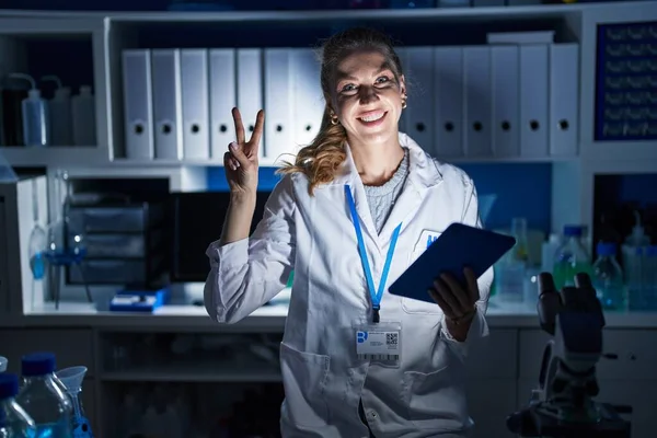 夜遅くに科学研究所で働く美しいブロンドの女性は 指で勝利サインをするカメラに顔をウィンクして笑顔で — ストック写真
