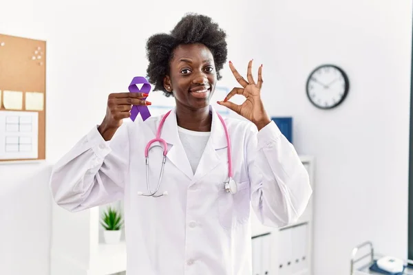 African doctor woman holding purple ribbon awareness at medical clinic doing ok sign with fingers, smiling friendly gesturing excellent symbol