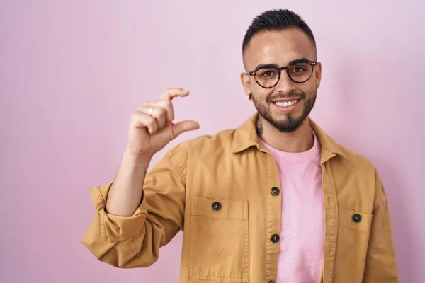ピンクの背景に立つ若いヒスパニック系の男が笑顔で 指を見てカメラで小さなサイズの看板を行う手で自信を持ってジェスチャー 概念を測定し — ストック写真