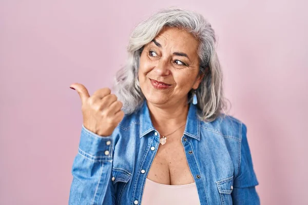 Middle age woman with grey hair standing over pink background smiling with happy face looking and pointing to the side with thumb up.