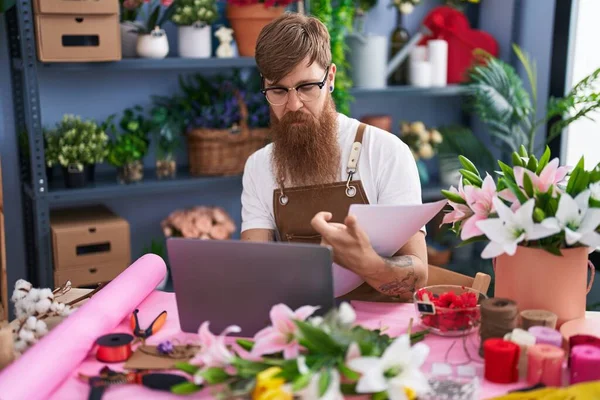 Young redhead man florist using laptop reading document at flower shop