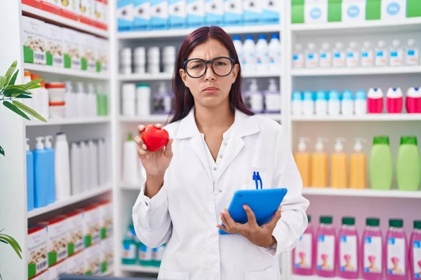 Young Hispanic Woman Working Pharmacy Drugstore Holding Red Heart Skeptic — Stock Photo, Image