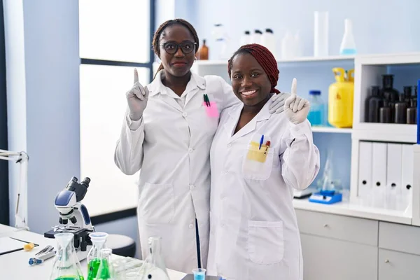 Two african women working at scientist laboratory surprised with an idea or question pointing finger with happy face, number one