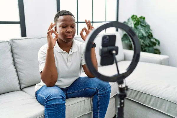 自宅でスマートフォンでVlogチュートリアルを記録した若いアフリカ人男性は 指で瞑想ジェスチャーを行う閉じて目でリラックスして笑顔 ヨガのコンセプト — ストック写真