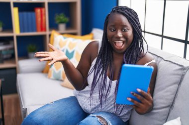 Young african woman using touchpad sitting on the sofa celebrating achievement with happy smile and winner expression with raised hand 