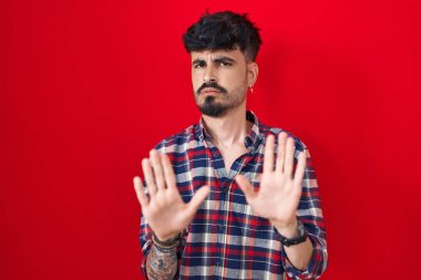Young hispanic man with beard standing over red background moving away hands palms showing refusal and denial with afraid and disgusting expression. stop and forbidden. 