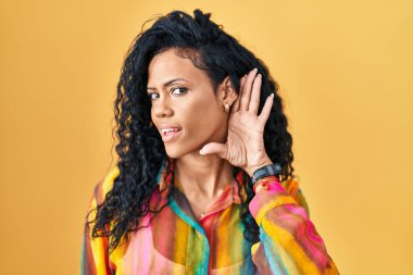 Middle age hispanic woman standing over yellow background smiling with hand over ear listening an hearing to rumor or gossip. deafness concept. 