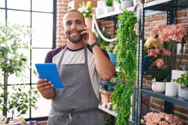 Young latin man florist talking on smartphone using touchpad at flower shop