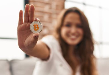 Young beautiful hispanic woman holding bitcoin sitting on sofa at home