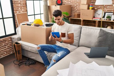 Yeni evindeki koltukta oturan dokunmatik ped kullanan İspanyol genç adam.