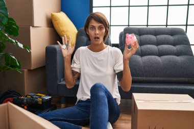 Brunette woman holding keys of new home and piggy bank afraid and shocked with surprise and amazed expression, fear and excited face. 