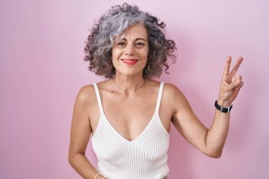 Pembe arka planda duran gri saçlı orta yaşlı kadın kameraya gülümseyen ve parmaklarıyla zafer işareti yapan. İki numara.. 