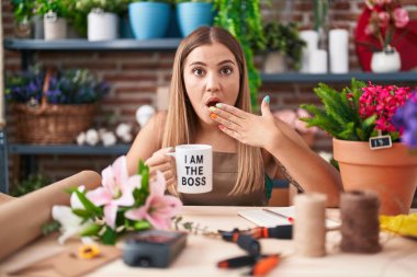 Young blonde woman working at florist shop holding i am the boss cup covering mouth with hand, shocked and afraid for mistake. surprised expression 