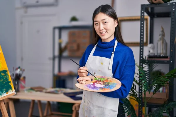 Chinese woman artist smiling confident holding paintbrush and palette at art studio