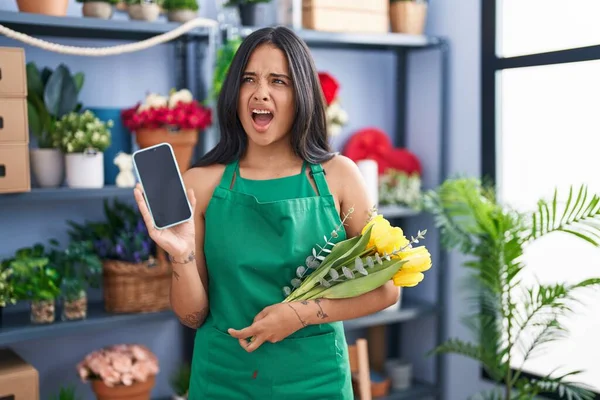 Brunette woman working at florist shop holding smartphone angry and mad screaming frustrated and furious, shouting with anger. rage and aggressive concept.