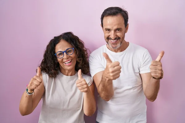 ピンクの背景の成功のサインの上に中年のパニックカップル一緒に手で積極的なジェスチャーを行うと 親指を笑顔と幸せ 陽気な表情と勝者のジェスチャー — ストック写真