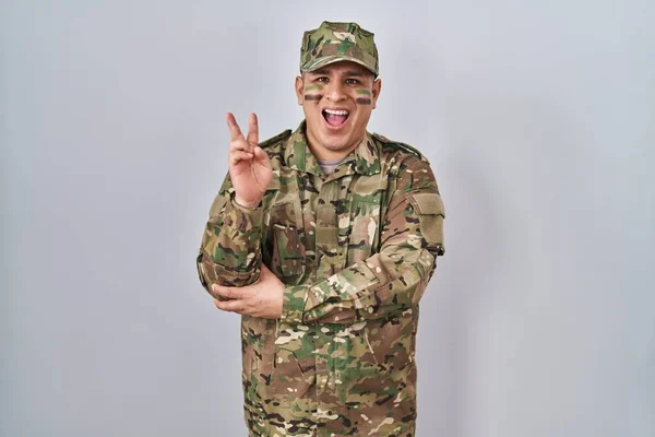 Hispanic young man wearing camouflage army uniform smiling with happy face winking at the camera doing victory sign. number two.