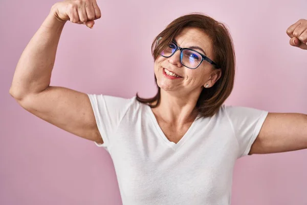 ピンクの背景の上に立つ中年のパニック女性は腕の筋肉を誇りに思って笑顔を見せる フィットネスのコンセプト — ストック写真