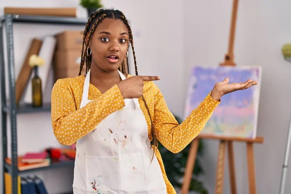 African american woman with braids at art studio amazed and smiling to the camera while presenting with hand and pointing with finger.