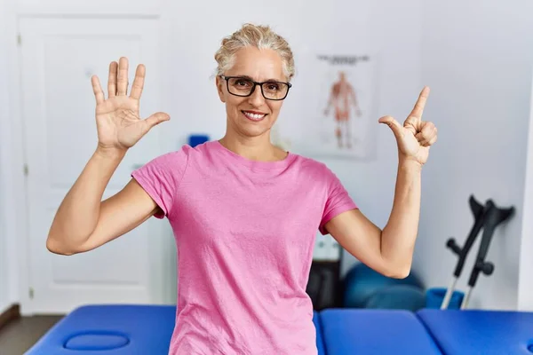 Middle age blonde woman at pain recovery clinic showing and pointing up with fingers number seven while smiling confident and happy.