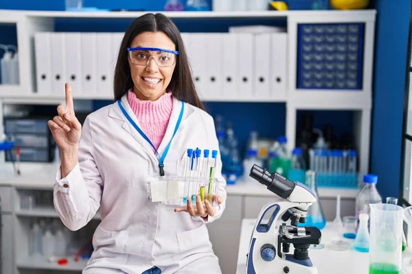 Young brunette woman working at scientist laboratory surprised with an idea or question pointing finger with happy face, number one