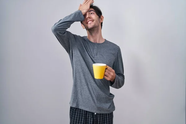 Young hispanic man wearing pajama drinking a cup of coffee surprised with hand on head for mistake, remember error. forgot, bad memory concept.