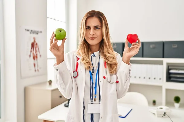 Young Blonde Doctor Woman Holding Heart Green Apple Skeptic Nervous — Stock Photo, Image