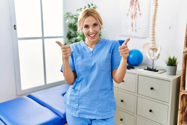Beautiful blonde physiotherapist woman working at pain recovery clinic success sign doing positive gesture with hand, thumbs up smiling and happy. cheerful expression and winner gesture.