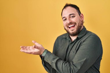 Plus size hispanic man with beard standing over yellow background pointing aside with hands open palms showing copy space, presenting advertisement smiling excited happy 