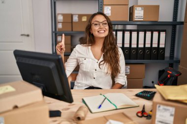 Young hispanic woman working at small business ecommerce showing and pointing up with finger number one while smiling confident and happy. 