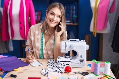 Young caucasian woman tailor talking on smartphone writing on notebook at sewing studio