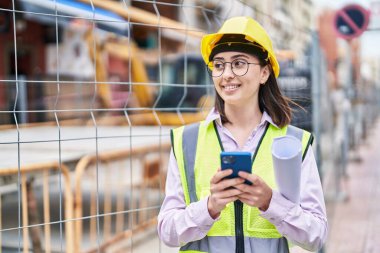 Young hispanic woman architect using smartphone holding blueprints at street