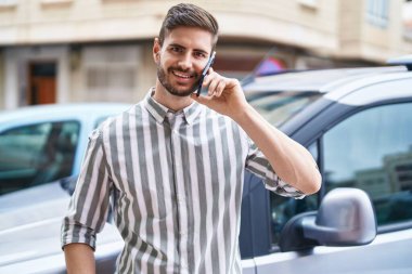 Beyaz bir adam akıllı telefondan konuşuyor. Sokakta arabaya yaslanıyor.